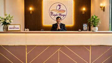 A reception desk with a sign reading 'The Purple Mango' and a 'RECEPTION' sign. There are two potted plants on either side of the desk.