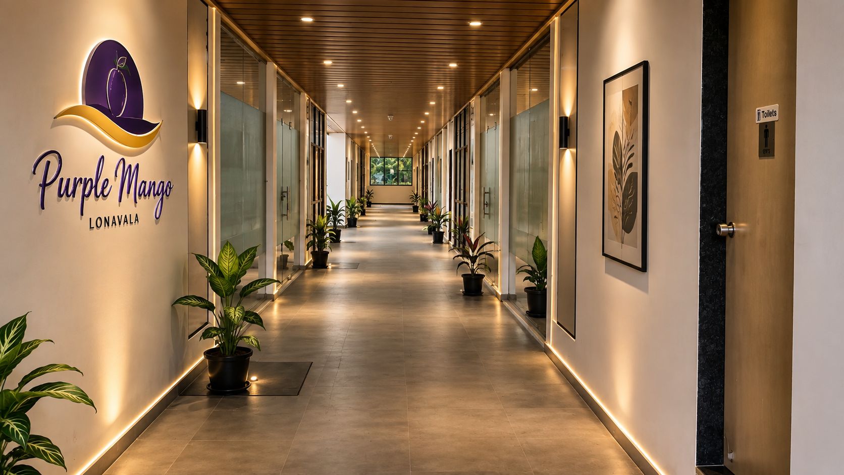 A hotel corridor with a sign for 'Purple Mango Lonavala,' potted plants, and a sign indicating toilets.