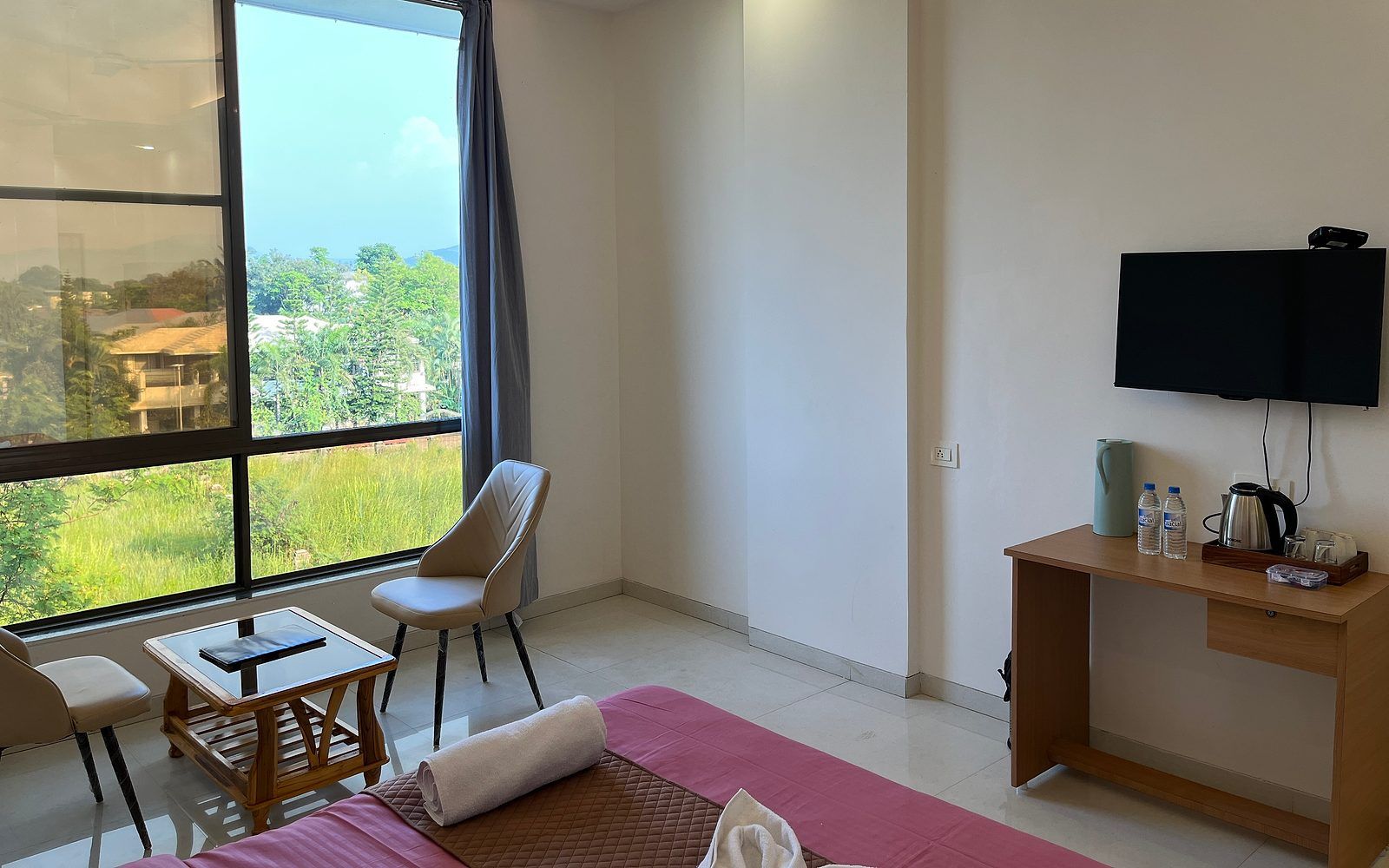 Habitación de hotel con cama, escritorio, silla, televisor y ventana con vista a follaje.