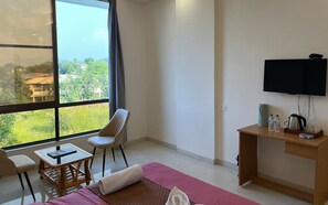 A hotel room with a bed, a desk, a chair, a TV, and a window with a view of greenery. - The Purple Mango - Pure Veg (Lonavala)