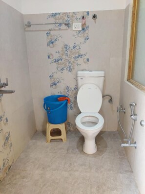 Un baño con inodoro, un cubo azul sobre un taburete y zona de ducha.