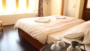 Una cama bien hecha con cabecera de madera, una mesita de noche con cafetera y vasos, y una ventana con cortinas.