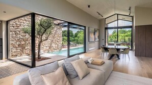 A modern living room with a large glass wall, a stone wall, a pool, and a dining area. - Villa Casa di Nova (Bale)