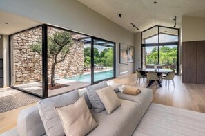 A modern living room with a large glass wall, a stone wall, a pool, and a dining area. - Villa Casa di Nova (Bale)