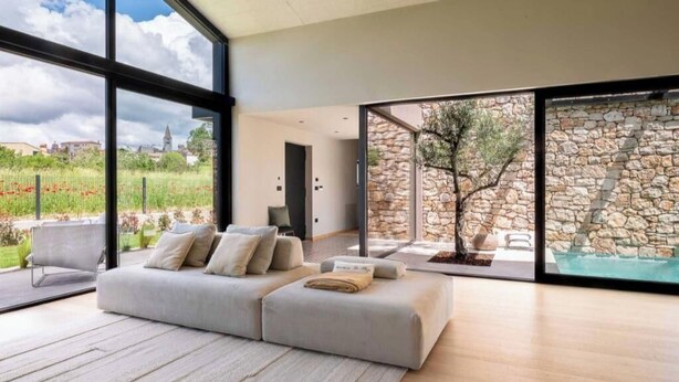 A modern living room with a large window, a grey sofa, and a stone wall. - Villa Casa di Nova (Bale)
