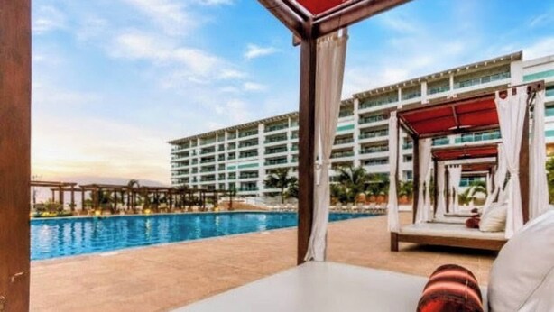 Zona de piscina de hotel con vistas a un edificio de varios pisos, un área de estar techada y un cielo despejado.