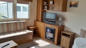 A compact living space with a sofa, a television, a small cabinet, and a heater. - Ty Mawr Holiday Park - Holiday Accommodation 21752 (Abergele)