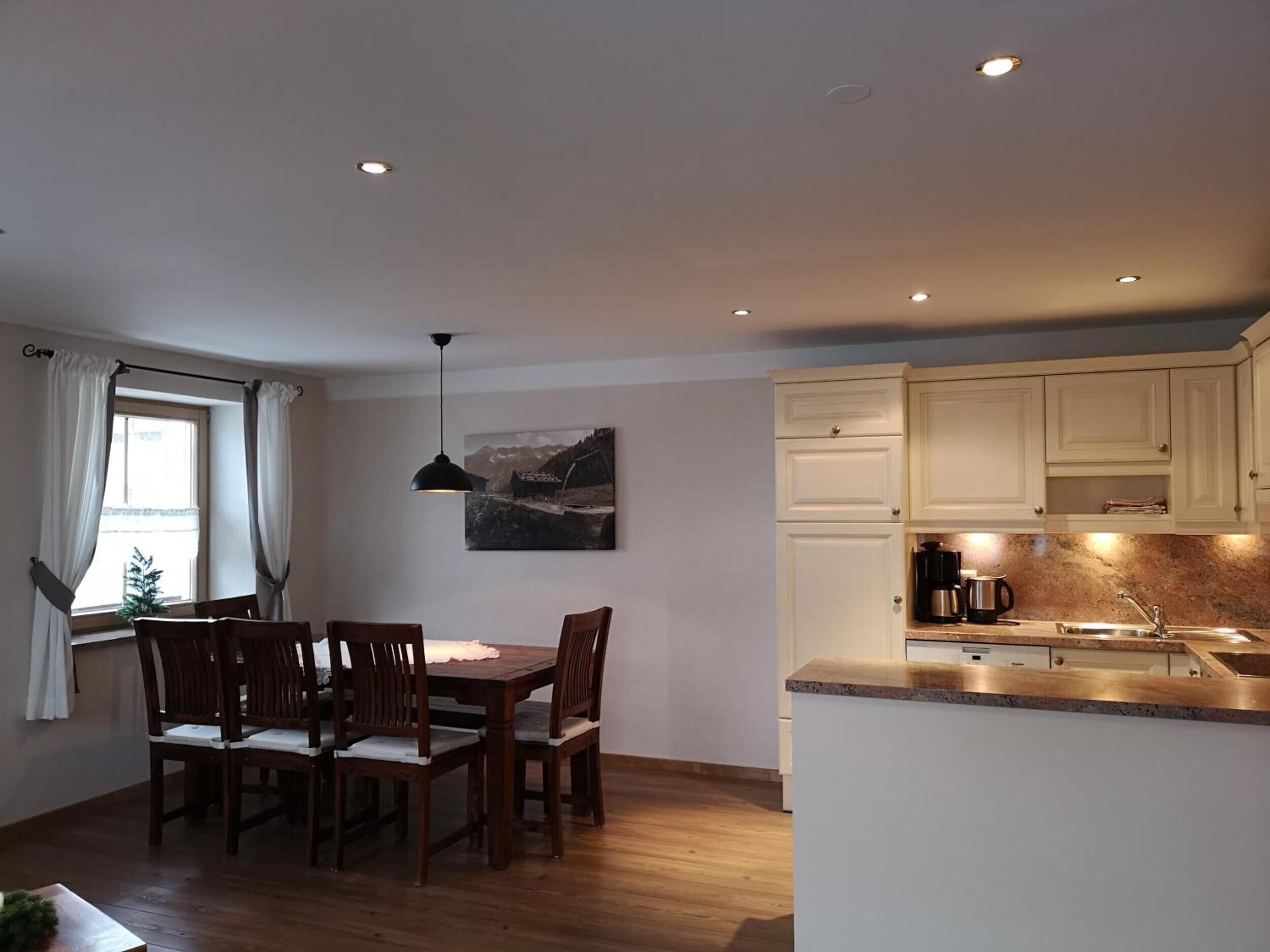 A dining area with a wooden table and chairs, a pendant light, and a painting on the wall.