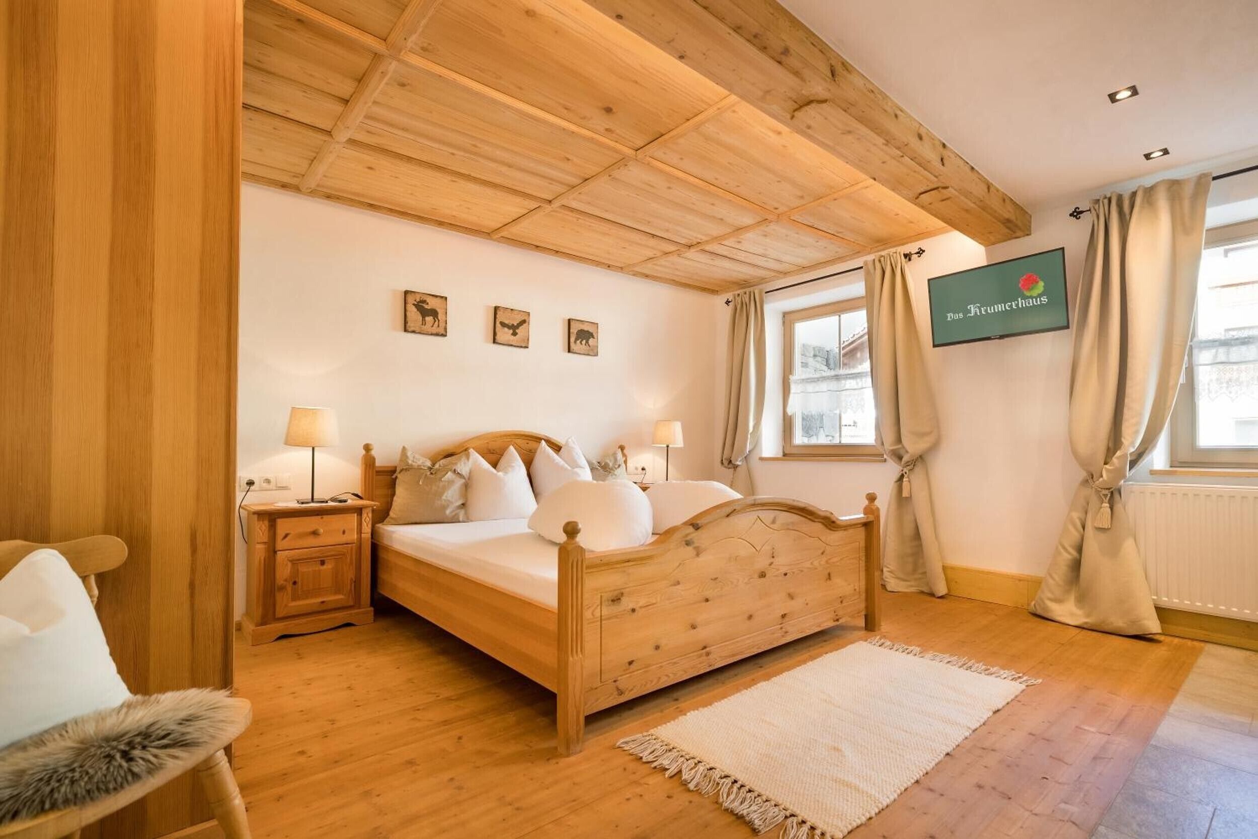 A wooden bed with a headboard, a bedside table with a lamp, a wall-mounted TV, and a window with curtains.