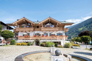A hotel with a traditional Alpine architectural style, featuring a central courtyard with a fountain, balconies with flower boxes, and a mountain backdrop.