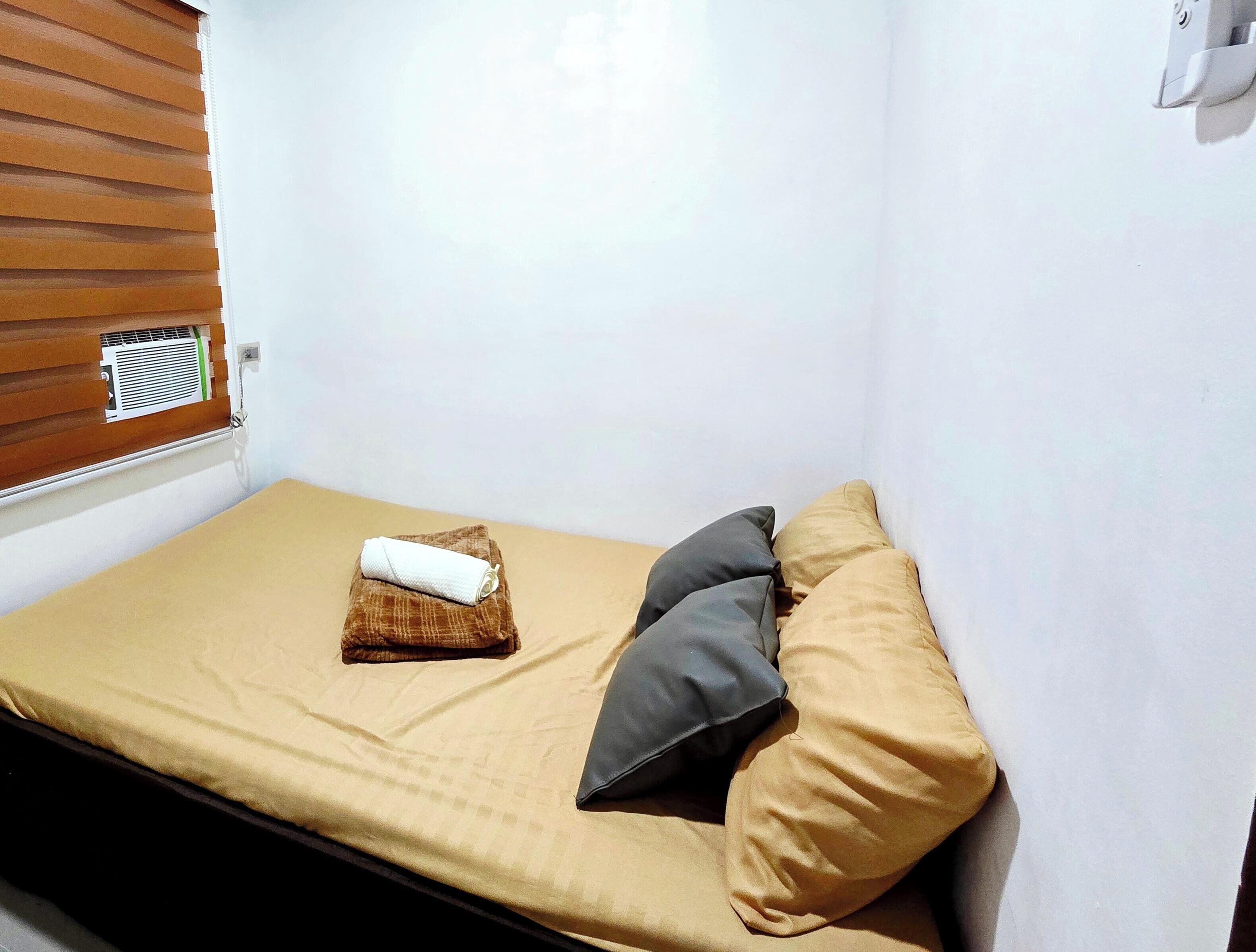A small, single-bed room with a beige mattress, a brown headboard, and a window with blinds.