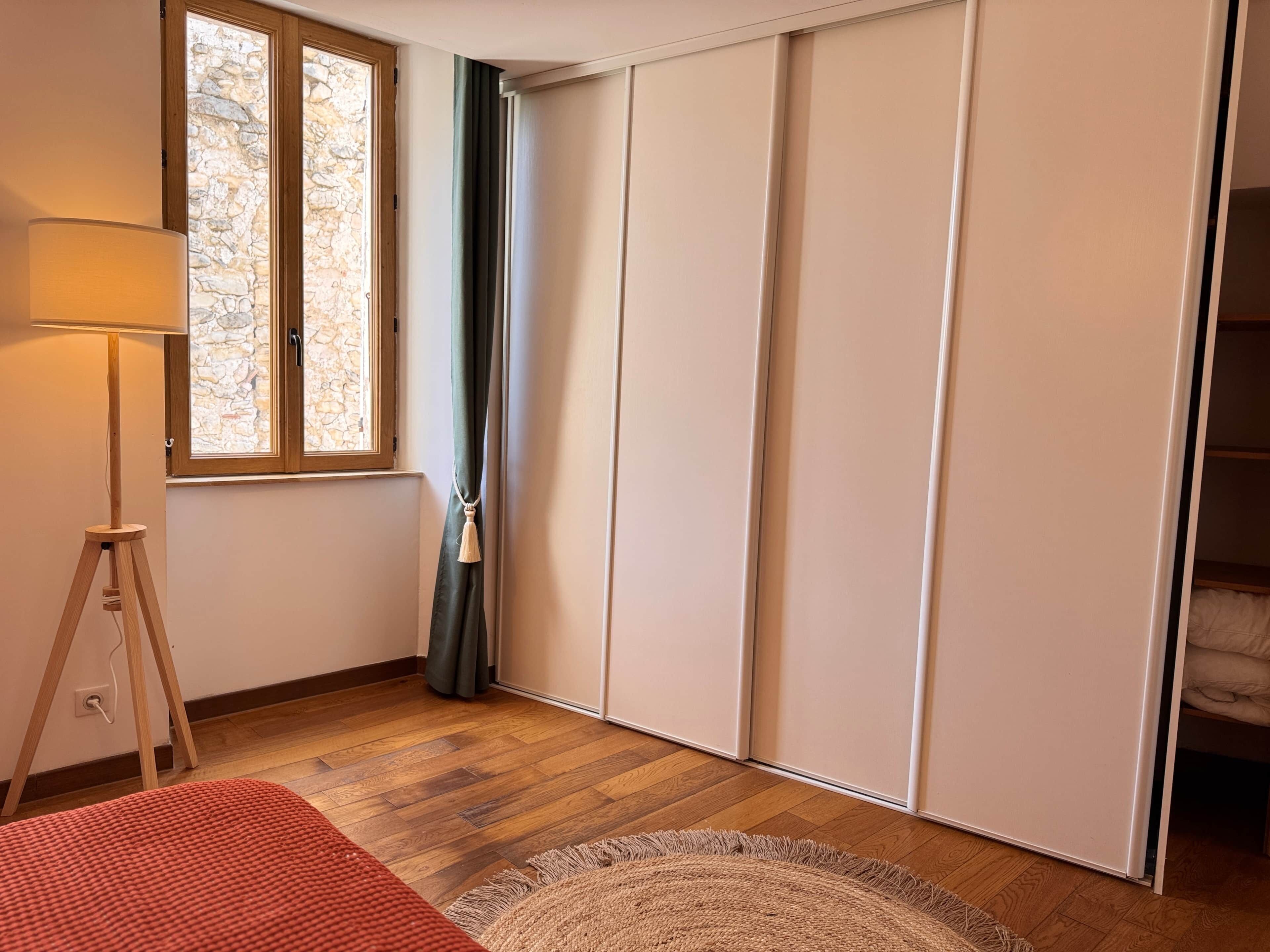 Ein Zimmer mit Holzboden, einer Lampe, einem fenster mit Vorhängen und einem Kleiderschrank.