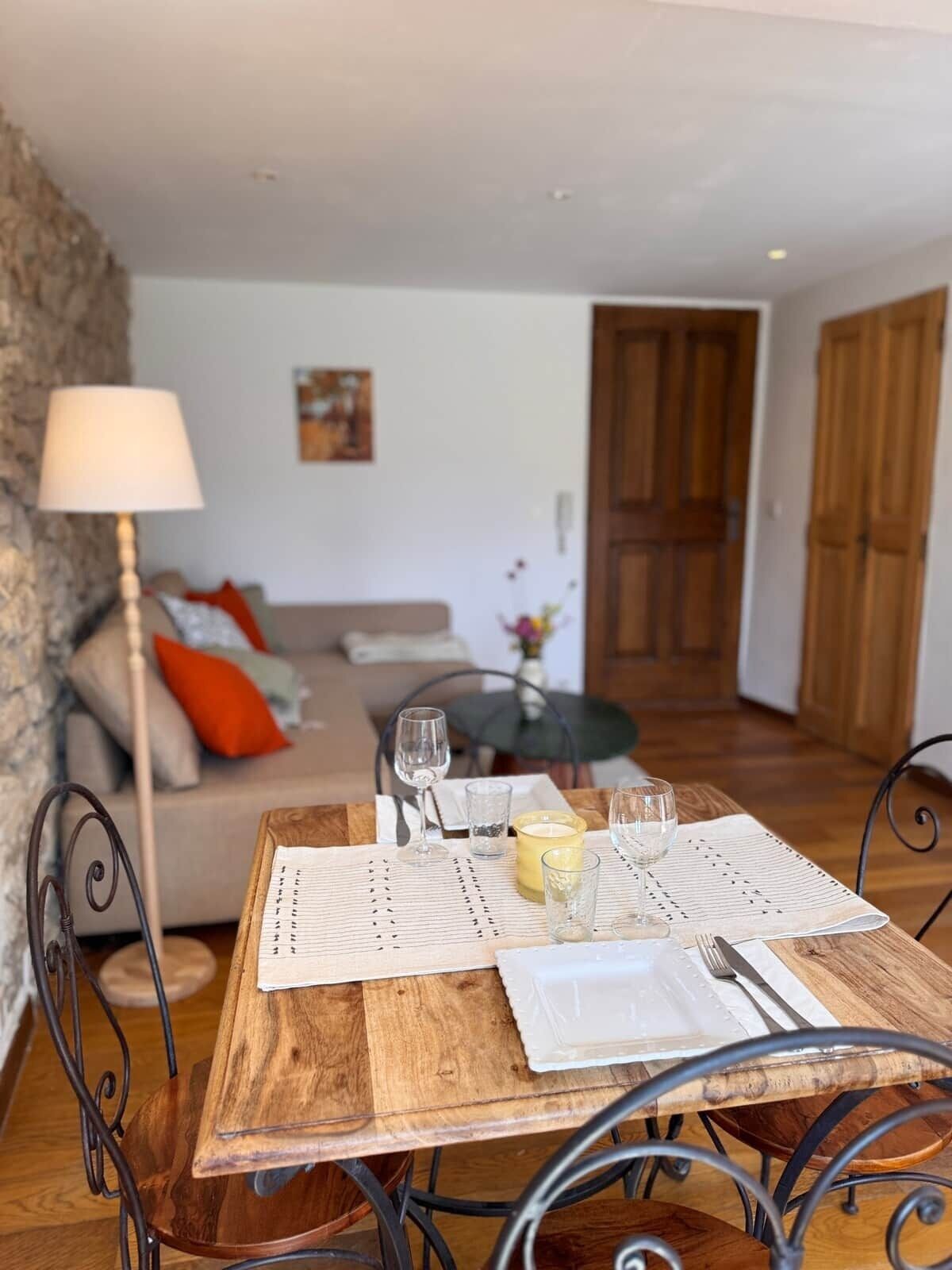 A dining table set with glasses, a candle, and a menu in a cozy living room with a sofa, a lamp, and a wooden door.