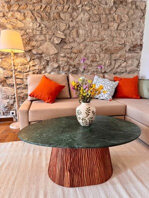 Uma sala de estar com parede de pedra, um sofá com almofadas laranja, uma mesa de centro redonda com um vaso de flores e um abajur.