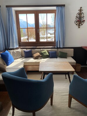 A living room with a sofa, two blue chairs, a wooden coffee table, and a window with curtains. - Nusshof Vacation Home for up to 9 people (Berchtesgaden)