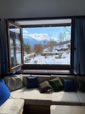 A room with a large window, a sofa with cushions, and a view of a snowy landscape with mountains. - Nusshof Vacation Home for up to 9 people (Berchtesgaden)