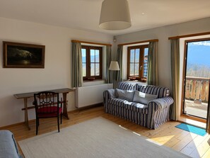 A room with a wooden floor, a striped sofa, a table with a chair, a radiator, and a window with curtains. - Nusshof Vacation Home for up to 9 people (Berchtesgaden)
