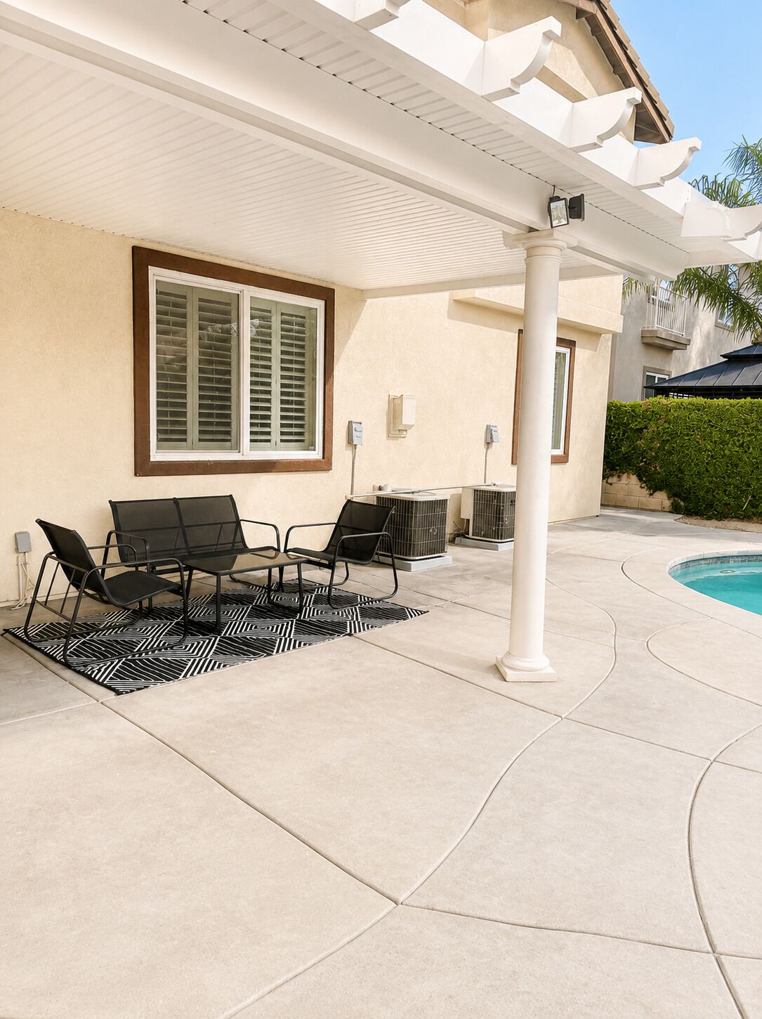 A covered patio area with outdoor furniture, a pool, and a building with a beige exterior.