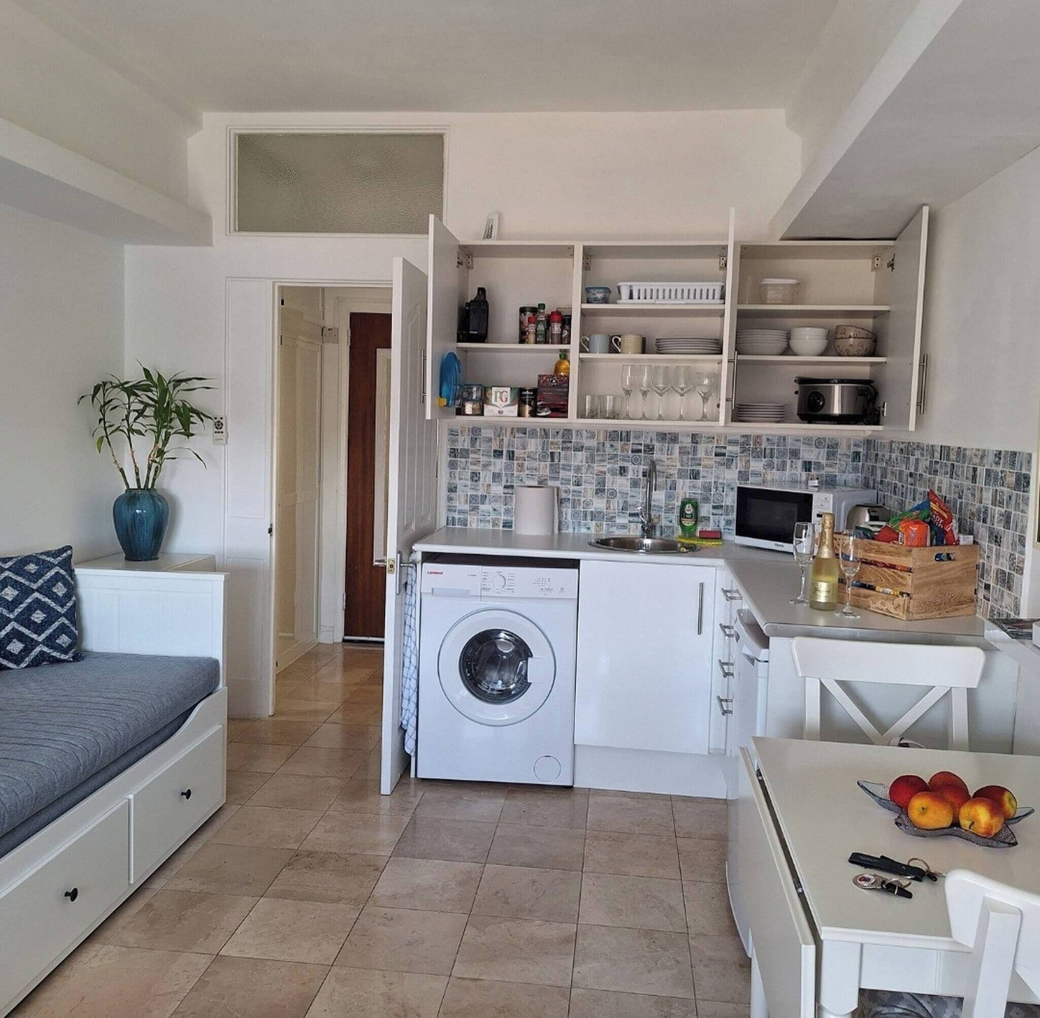 Une cuisine compacte équipée d’un lave-linge, d’une table à manger avec des fruits et d’un lit agrémenté d’un coussin à motifs.