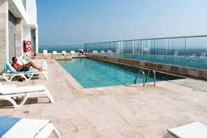 Ein Pool auf dem Dach mit klarem, blauem Wasser, weißen Liegestühlen und einem Glasgeländer.