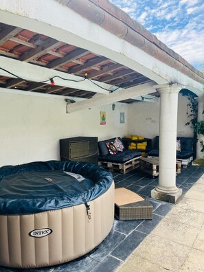 Ett takförsett utomhusområde med sittplatser, ett bubbelbad, möbler i rotting och en pelare.