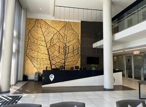 Un moderno vestíbulo de hotel con un mostrador de recepción, una gran obra de arte abstracta en la pared y una escalera que conduce a un nivel superior.