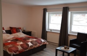 A hotel room with a bed, a small table, a chair, a window with curtains, and a radiator. - Vrbo Property (Altenau)