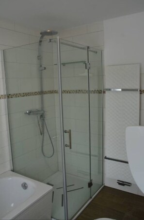 Een glazen douchecabine met handdouche, een witte wastafel en een toilet in een betegelde badkamer.