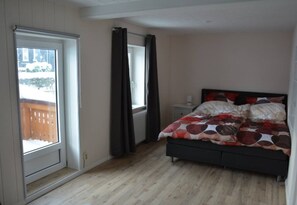 Een slaapkamer met een bed, een raam met gordijnen, een nachtkastje en uitzicht op een houten terras.