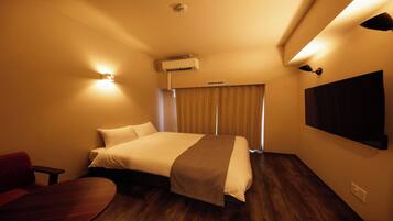 A hotel room with a bed, a desk, a chair, an air conditioning unit, a wall-mounted TV, and a window with curtains.