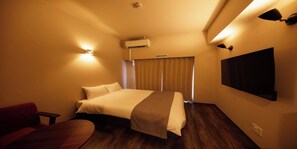 A hotel room with a bed, a desk, a chair, an air conditioning unit, a wall-mounted TV, and a window with curtains.
