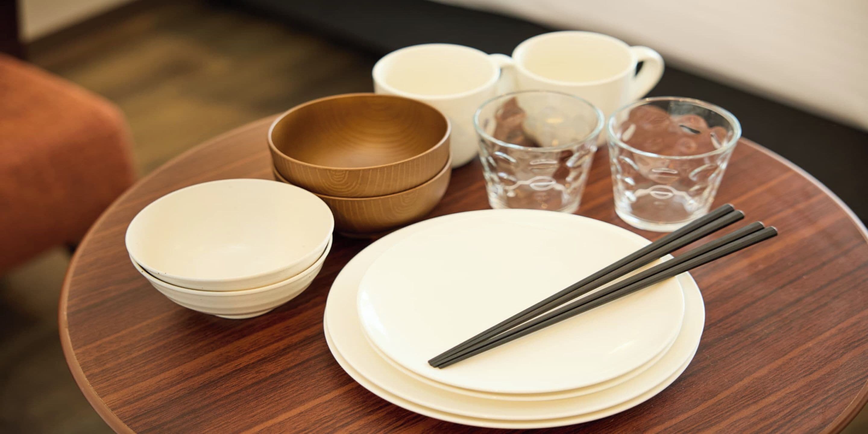 A round wooden table with stacked white plates, a pair of chopsticks, a brown bowl, and a glass of water.