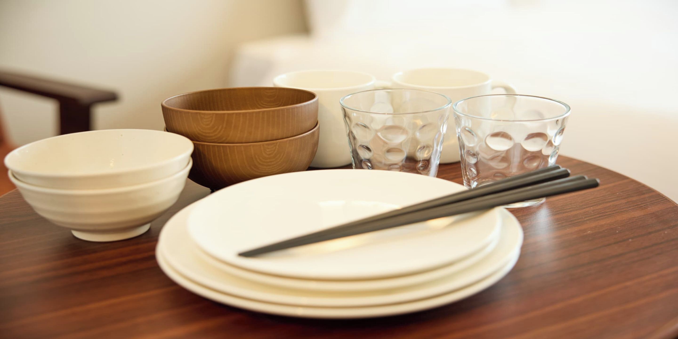 Una mesa de comedor con platos blancos apilados, un cuenco de madera y dos vasos de vidrio.