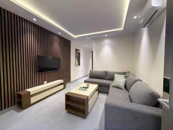A modern living room with a flat-screen TV, a grey sofa, a wooden coffee table, and a wall-mounted air conditioner. - 103 JRE comfortable and accessible retreat (Amman)