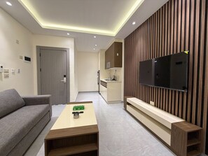 A modern living room with a sofa, a coffee table, a TV mounted on a wooden panel wall, a kitchen area in the background, and recessed ceiling lighting. - 103 JRE comfortable and accessible retreat (Amman)