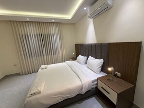Een moderne hotelkamer met een groot bed, een houten nachtkastje, een airconditioning en een raam met dunne gordijnen.
