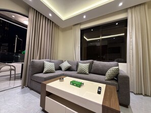 A modern living room with a grey sectional sofa, patterned cushions, a wooden coffee table, and a large window with curtains. - 201 JRE apartment with stunning boulevar (Amman)
