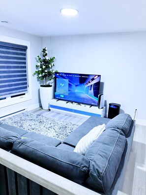 Ein modernes Zimmer mit einem großen Flachbildfernseher, einer Couch, einer Topfpflanze und einem Fenster mit Jalousien.