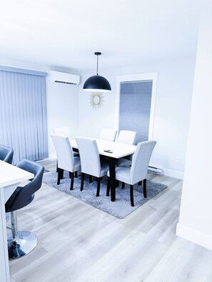 A modern dining area with a round table, six chairs, a black pendant light, and a white air conditioning unit. - Luxury 3-Bedroom Full House Near Magnetic Hill (Moncton)