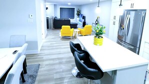 Una cocina moderna con un refrigerador de acero inoxidable, un comedor con mesa y sillas blancas, y un sofá amarillo brillante al fondo.