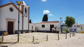 Un patio empedrado con un edificio blanco que tiene una torre del reloj, un caballo y un farol.