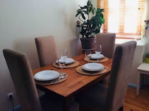 A dining table set with plates, glasses, and cutlery, surrounded by chairs, with a potted plant and a window with blinds in the background.