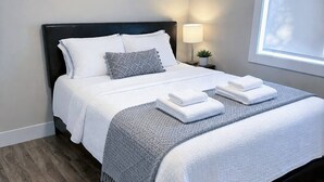 A neatly made bed with white linens and a gray throw, a bedside lamp, and a window with blinds. - Modern 3-Bedroom Retreat Near Moncton Hospital and Downtown (Moncton)