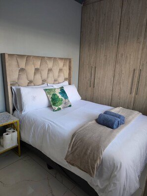 Een netjes opgemaakt bed met een gestoffeerd hoofdbord, een nachtkastje met een rol toiletpapier en een garderobe met twee deuren.