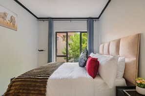A bedroom with a bed, a window with curtains, a painting on the wall, and a view of greenery outside. - Zalari Loft B, Nature-Inspired 2 Bed Loft Paulshof (Sandton)