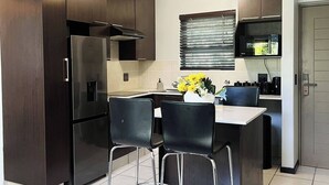 A compact kitchen with dark cabinets, a stainless steel refrigerator, a microwave, and a small dining area with two chairs. - Zalari Luxe Loft B, Nature 1-Bed Loft, Paulshof (Sandton)