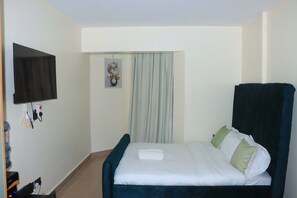 A neatly made bed with pillows, a flat-screen TV mounted on the wall, and a power outlet with a water bottle on the floor. - Skyline Luxury in South B | Stylish 11th Floor Retreat Near JKIA- Wynloft stays. (Nairobi)