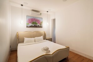 Uma cama bem arrumada com lençóis brancos e uma toalha enrolada, um quadro de cerejeiras em flor e um ar-condicionado no teto.
