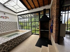 Un espace spa doté d’un jacuzzi à jets, protégé par des parois en verre, d’une cheminée en briques et de grandes fenêtres offrant une vue sur la verdure.