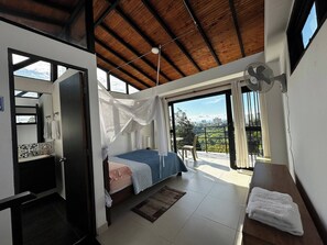 Une chambre à coucher avec un lit, une table de chevet, un ventilateur et une vue sur la ville grâce à de grandes fenêtres.